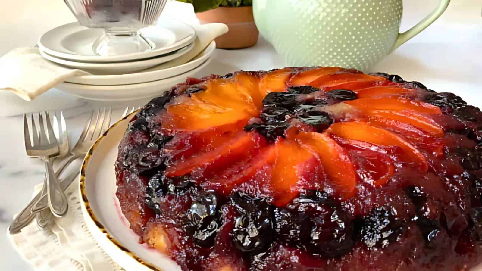 A fruit upside-down cake with glazed peach and cherry slices on top sits on a plate next to forks, plates, and a green teapot.
