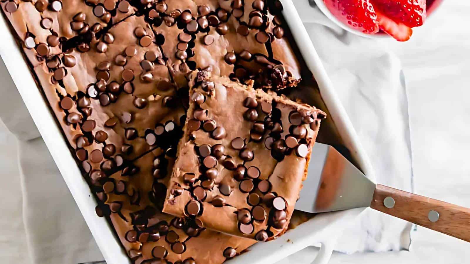 A baking dish of chocolate chip brownies with a spatula lifting one piece, next to sliced strawberries on a white surface.