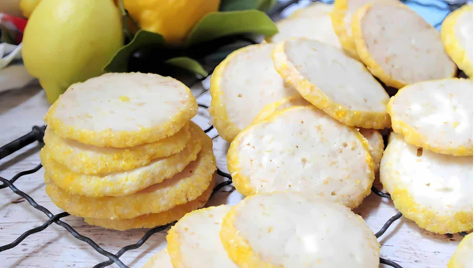 Round lemon cookies with a glazed topping are stacked and scattered on a cooling rack, with fresh lemons in the background.