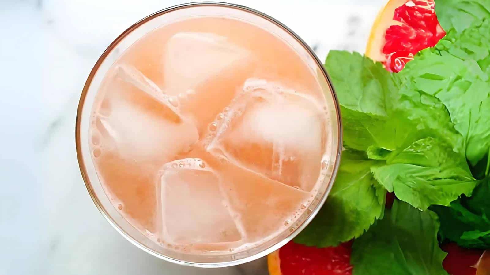 A glass filled with iced pink beverage, garnished with fresh mint leaves and slices of grapefruit on the side.