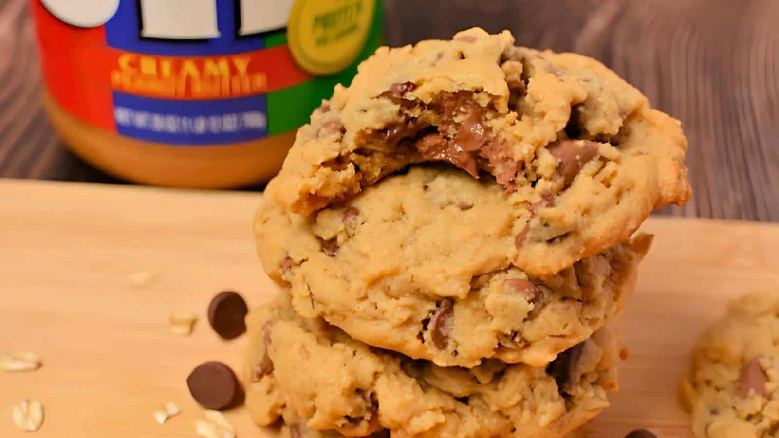 A stack of chocolate chip cookies sits on a wooden board next to a jar of creamy peanut butter and scattered chocolate chips.