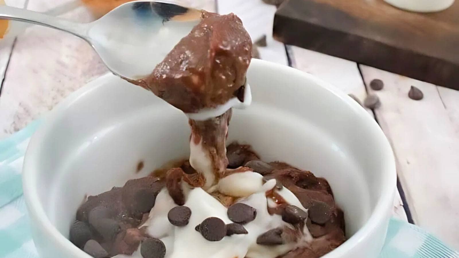 A spoon lifts a portion of chocolate dessert with cream and chocolate chips from a white bowl on a wooden table.
