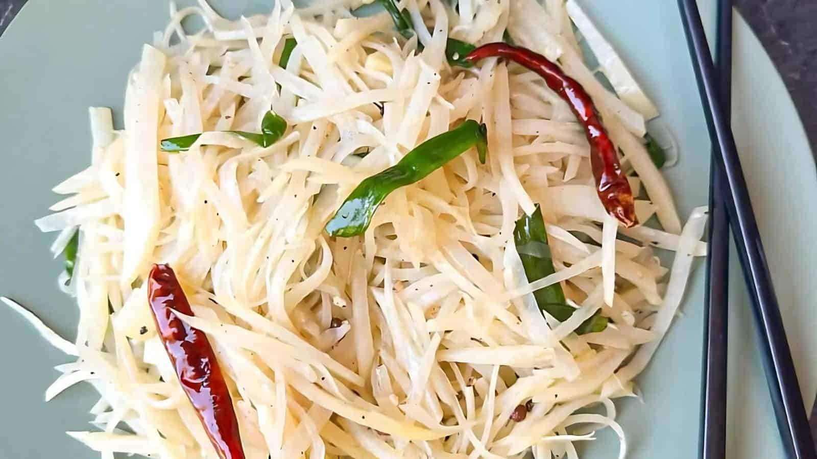 A plate of stir-fried shredded potatoes garnished with green onions and dried red chilies, served with black chopsticks.