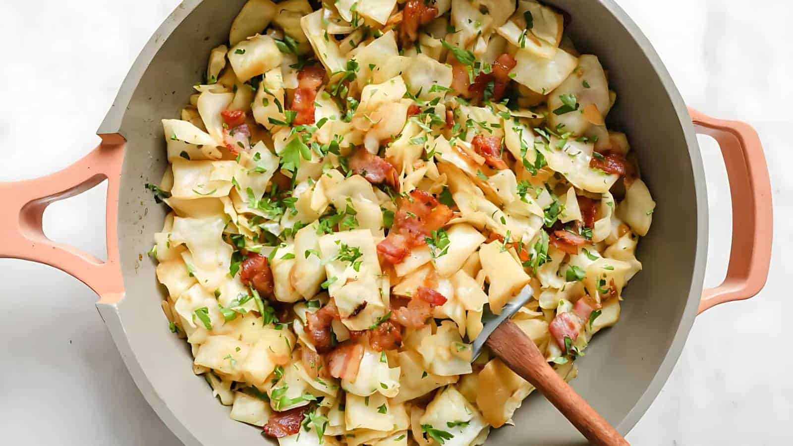 A pan filled with cooked cabbage, wide noodles, bacon pieces, and chopped parsley, with a wooden spoon resting inside.