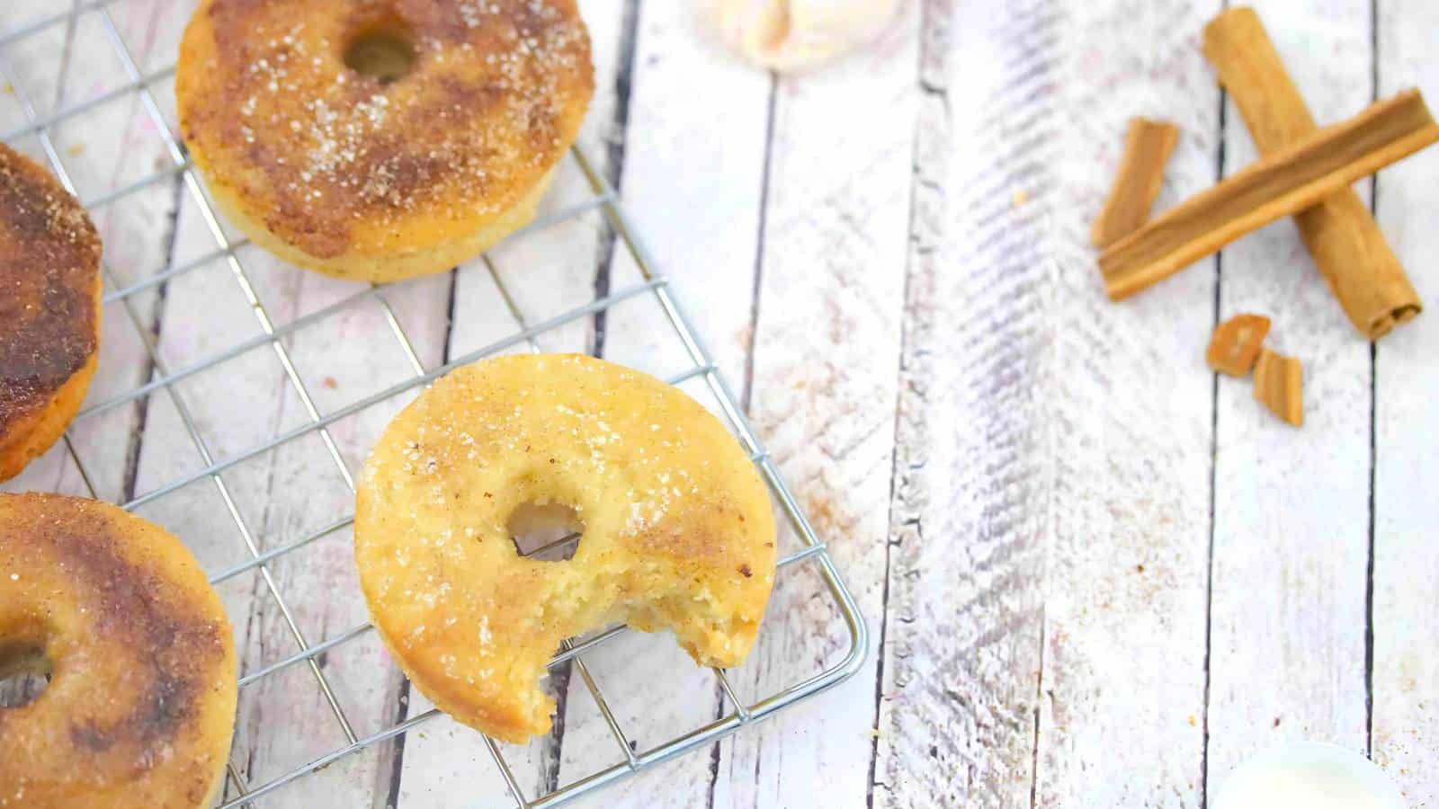 Three baked donuts on a cooling rack, one with a bite taken out, next to cinnamon sticks on a white wooden surface.