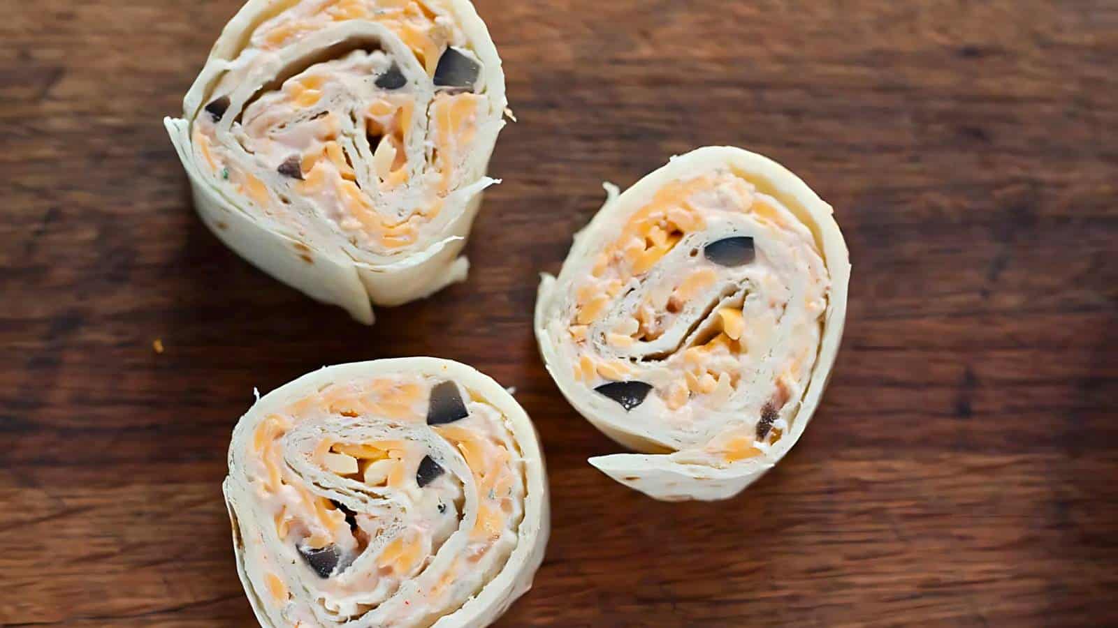Three tortilla pinwheels filled with a mixture of shredded cheese, chopped black olives, and a creamy spread are displayed on a wooden surface.