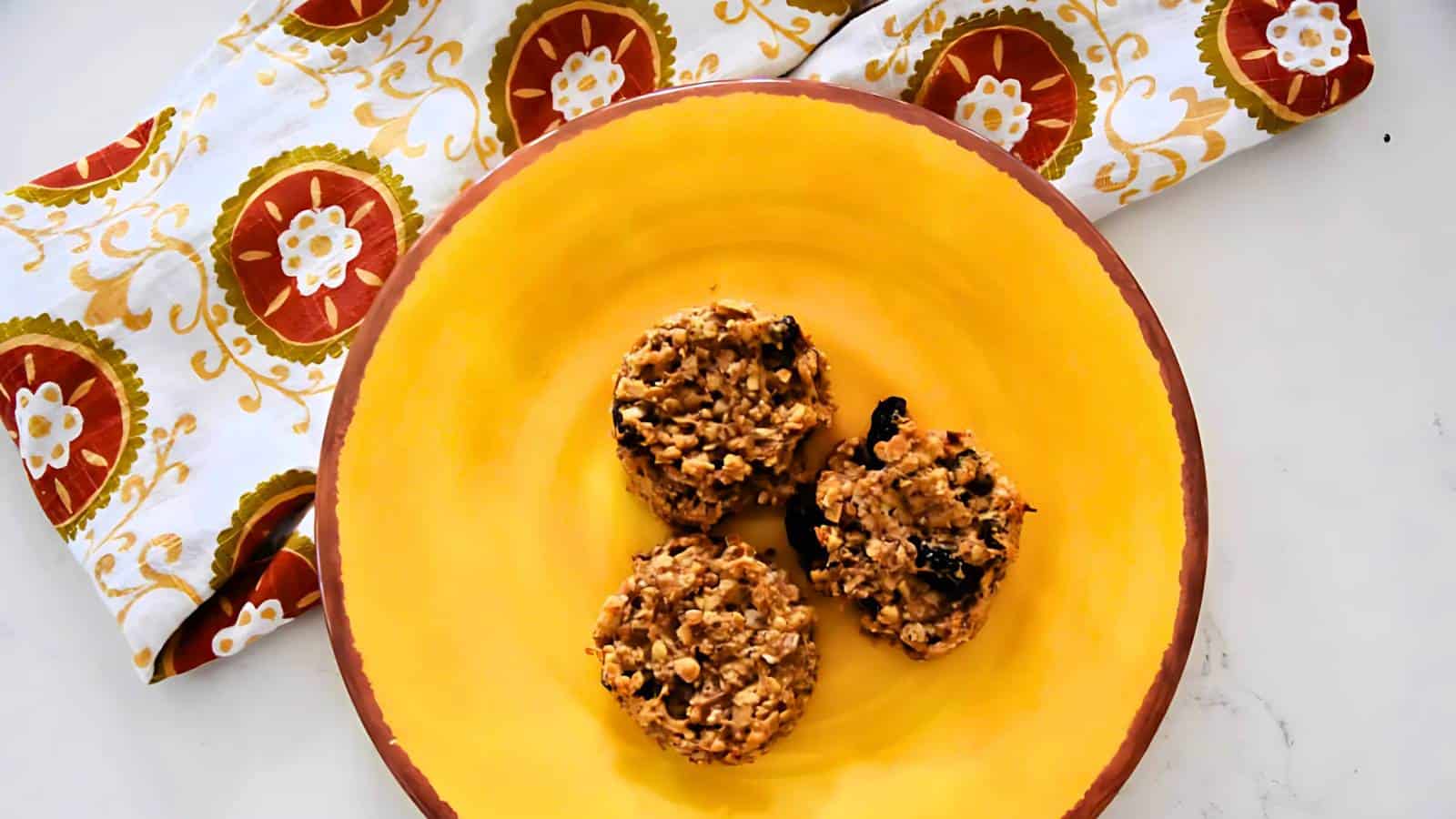 Three oatmeal cookies are arranged on a yellow plate, with a patterned cloth featuring red and green circular designs in the background.