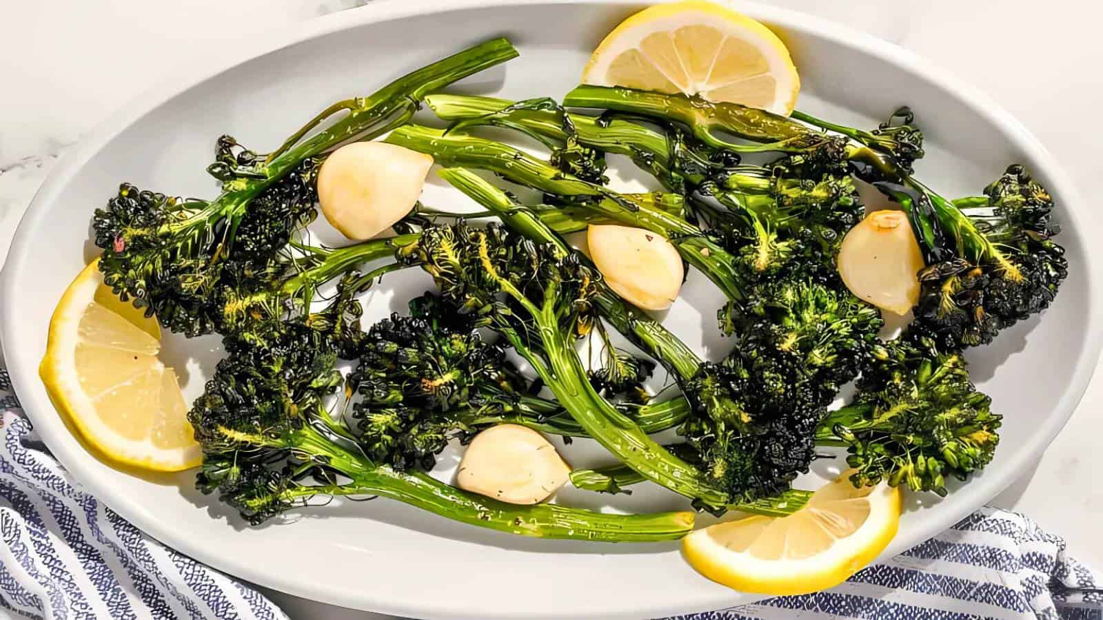 Roasted broccolini with whole garlic cloves and lemon slices served on a white oval plate, with a striped cloth partially visible.