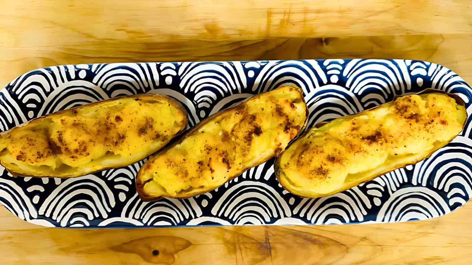 Three twice-baked potatoes topped with golden cheese are arranged in an oval dish with a blue and white patterned design, set on a wooden surface.
