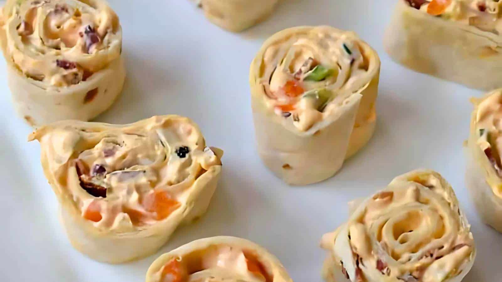 Close-up of several tortilla pinwheels filled with a creamy mixture and small pieces of vegetables and other ingredients, arranged on a white surface.