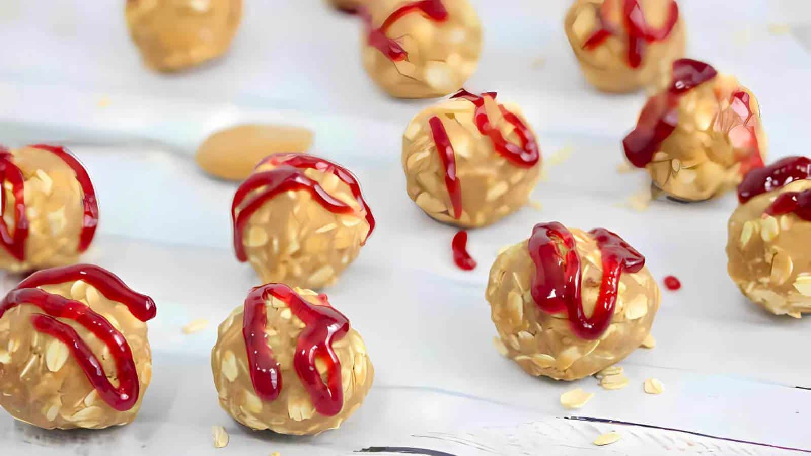 Round oat and peanut butter balls are drizzled with red jelly, arranged on a white surface with scattered oats.