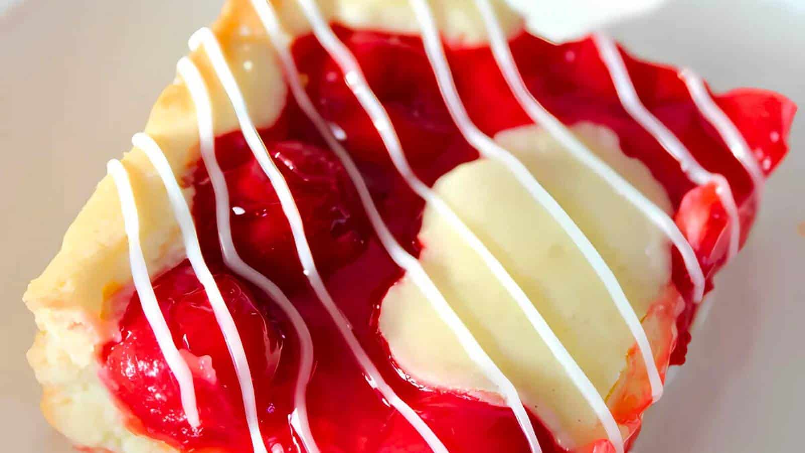 A close-up of a cherry dessert bar topped with cherries, icing drizzle, and a light pastry crust on a white plate.