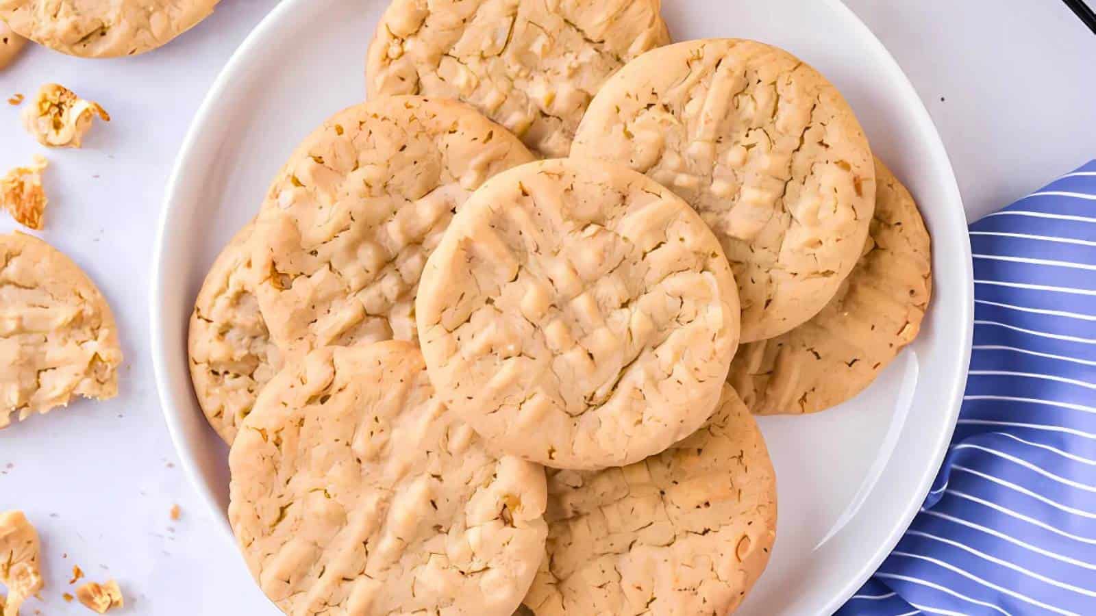 A plate of round, light brown peanut butter cookies with crisscross fork marks on top, with a few broken cookie pieces nearby.
