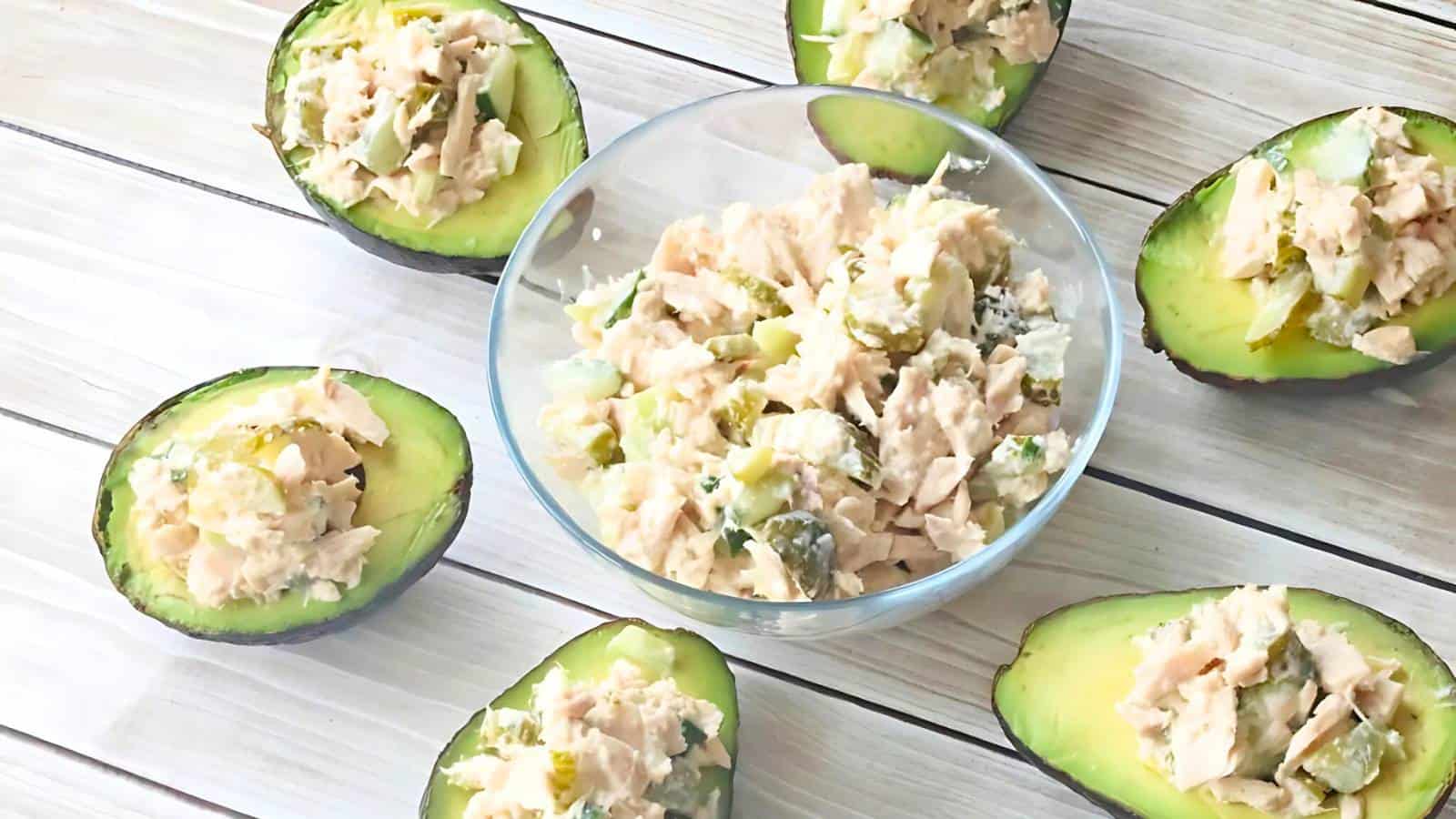 A glass bowl of tuna salad is surrounded by halved avocados filled with the same tuna salad, all arranged on a light wooden surface.