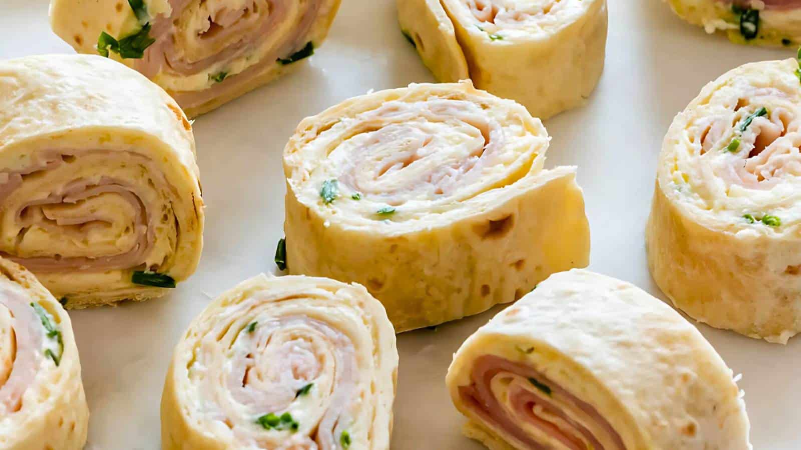 Close-up of sliced pinwheel appetizers made with tortillas, deli meat, cream cheese, and herbs on a white surface.