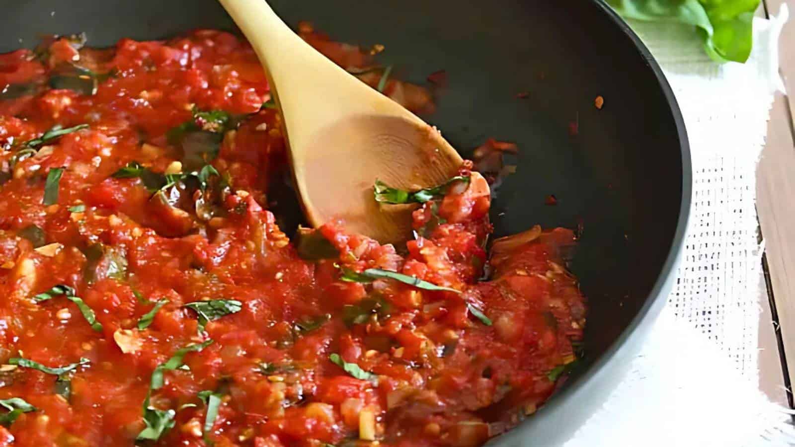 A wooden spoon stirs a pan of chunky tomato sauce with chopped herbs, likely basil, on a stovetop.