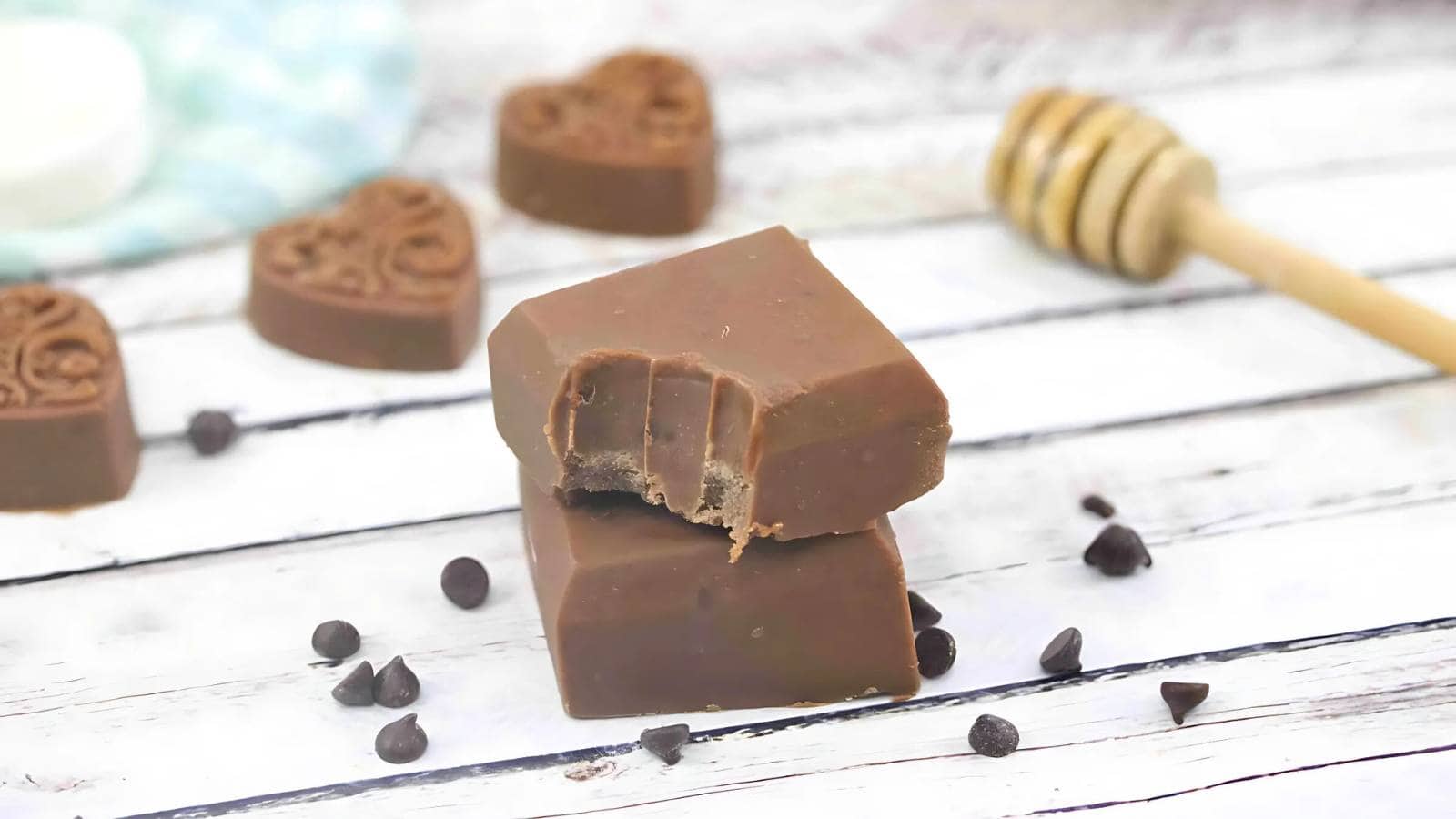 Two pieces of chocolate, one with bite marks, are stacked on a white wooden surface, surrounded by chocolate chips, heart-shaped chocolates, and a honey dipper.