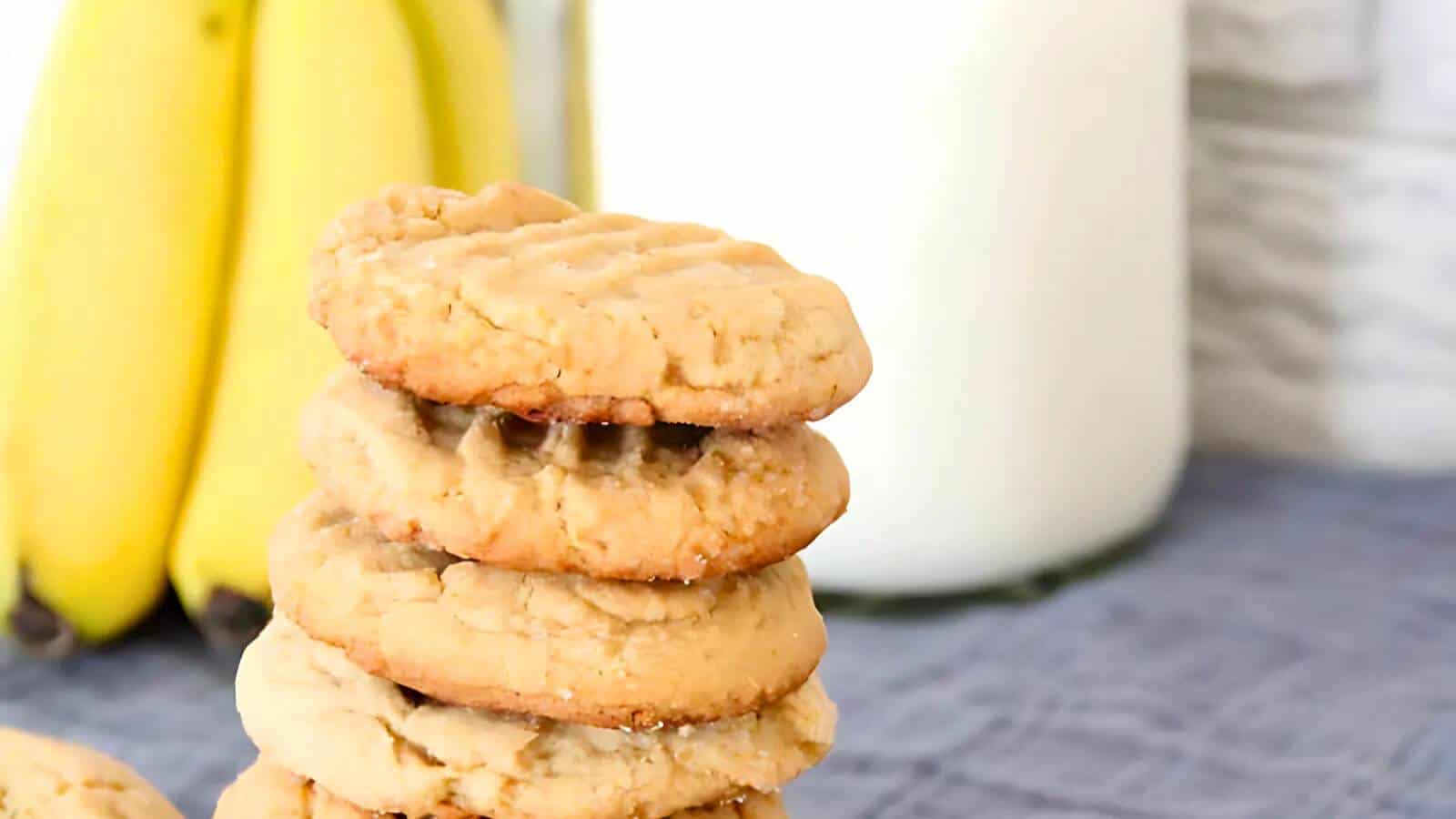 A stack of peanut butter cookies is placed in front of a bunch of bananas and a glass of milk on a gray cloth.