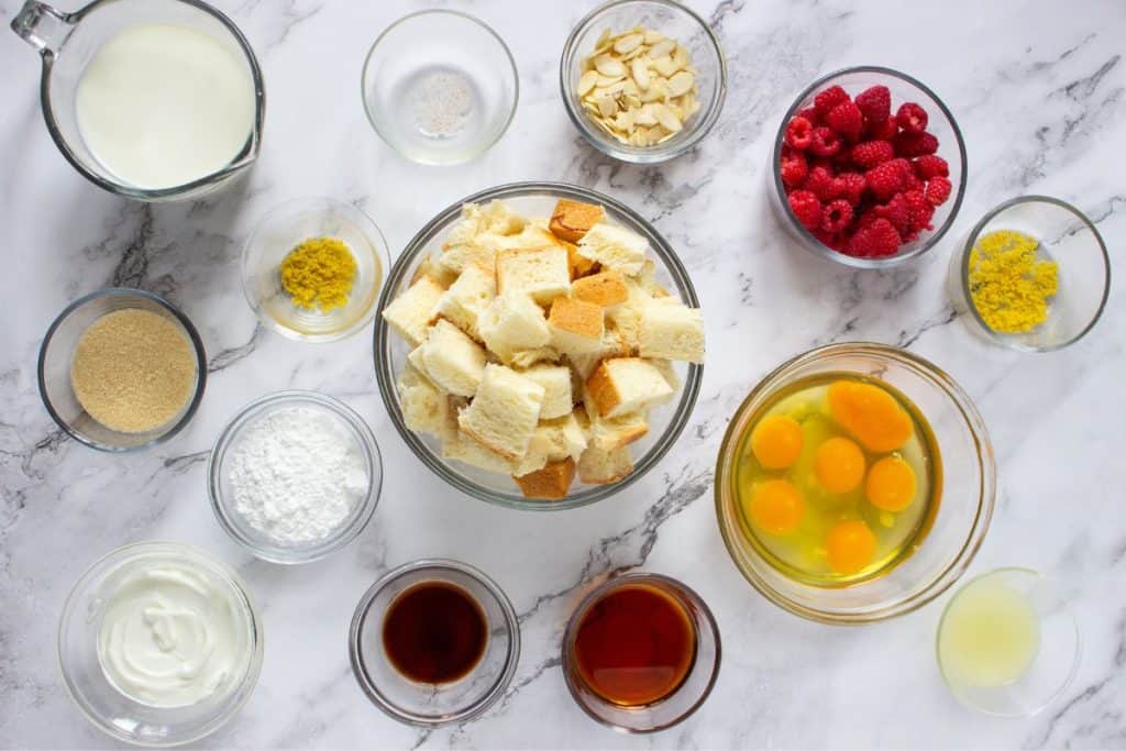 Various baking ingredients in glass bowls, including cubed bread, eggs, raspberries, milk, yogurt, almond slices, and assorted powders, are arranged on a marble surface.