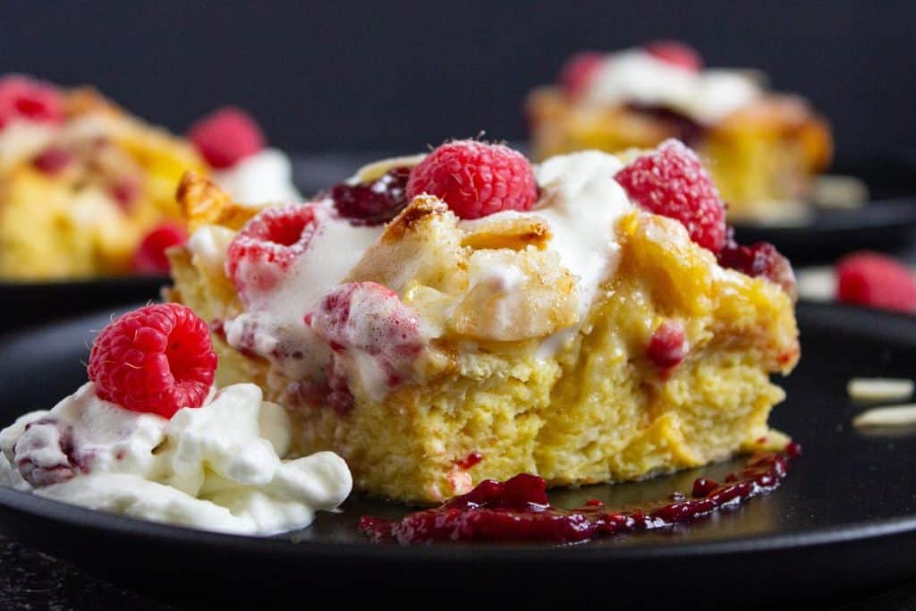 A slice of Brioche French Toast Casserole with whipped cream and fresh raspberries on a black plate, with a drizzle of red sauce nearby.