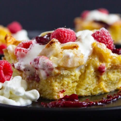 A slice of Brioche French Toast Casserole with whipped cream and fresh raspberries on a black plate, with a drizzle of red sauce nearby.
