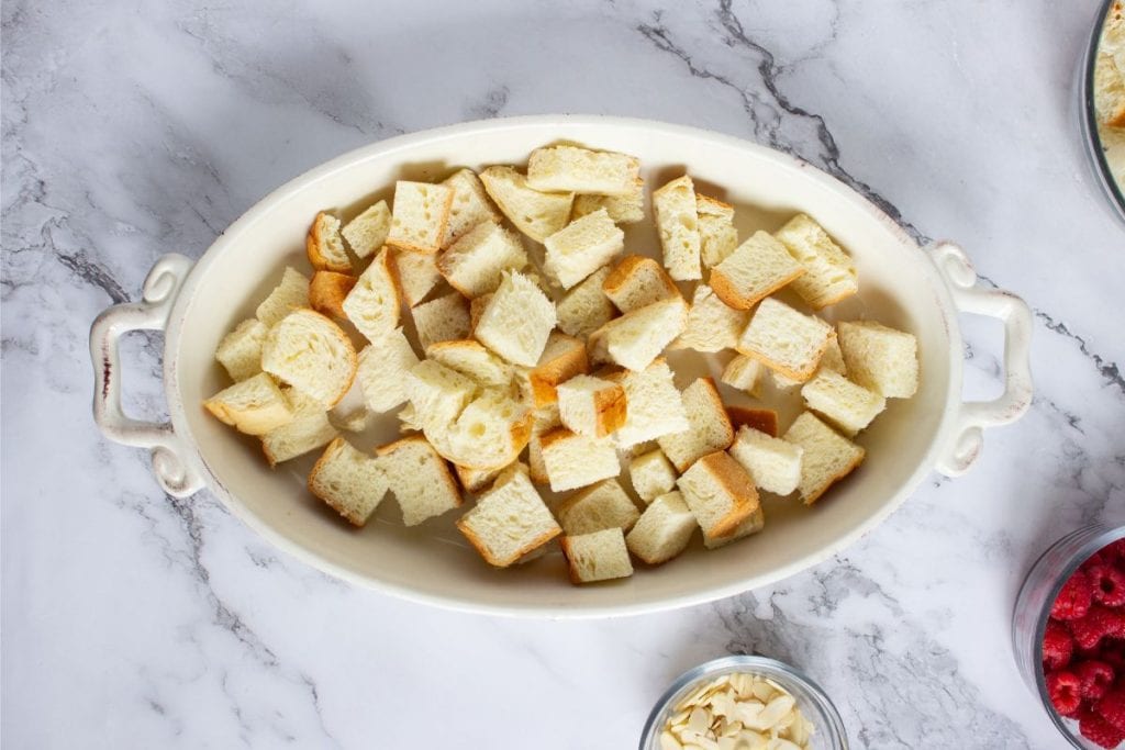 Oval casserole dish filled with cubed bread pieces on a marble countertop, with bowls of sliced almonds and raspberries nearby.