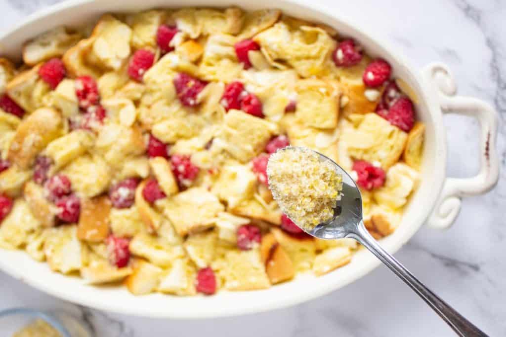 A spoon holds a portion of crumb topping above a white dish filled with a baked bread pudding dessert with raspberries.
