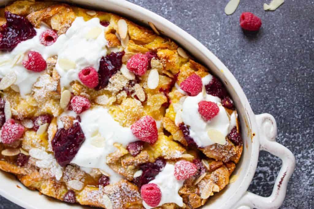 Baked Brioche French Toast Casserole in a white dish, topped with whipped cream, raspberries, sliced almonds, powdered sugar, and fruit preserves.