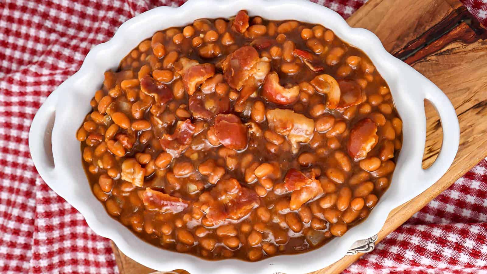A white dish filled with baked beans mixed with pieces of bacon, placed on a wooden board over a red and white checkered cloth.