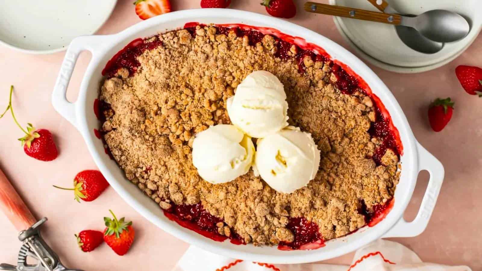 An oval dish filled with strawberry crumble topped with three scoops of vanilla ice cream, surrounded by fresh strawberries and dessert plates.