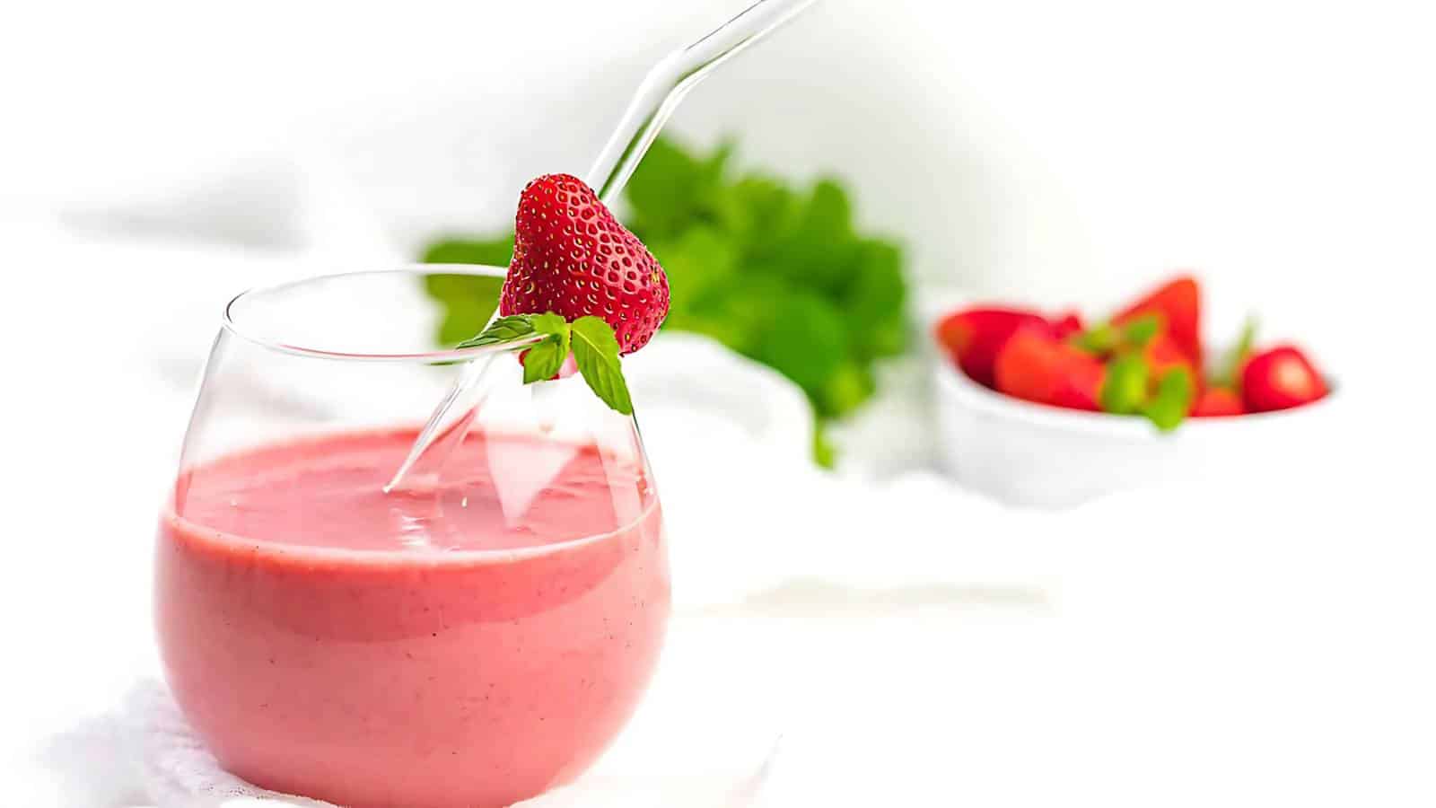 A glass of pink strawberry smoothie with a straw and a whole strawberry garnish, with a bowl of strawberries and green leaves in the background.