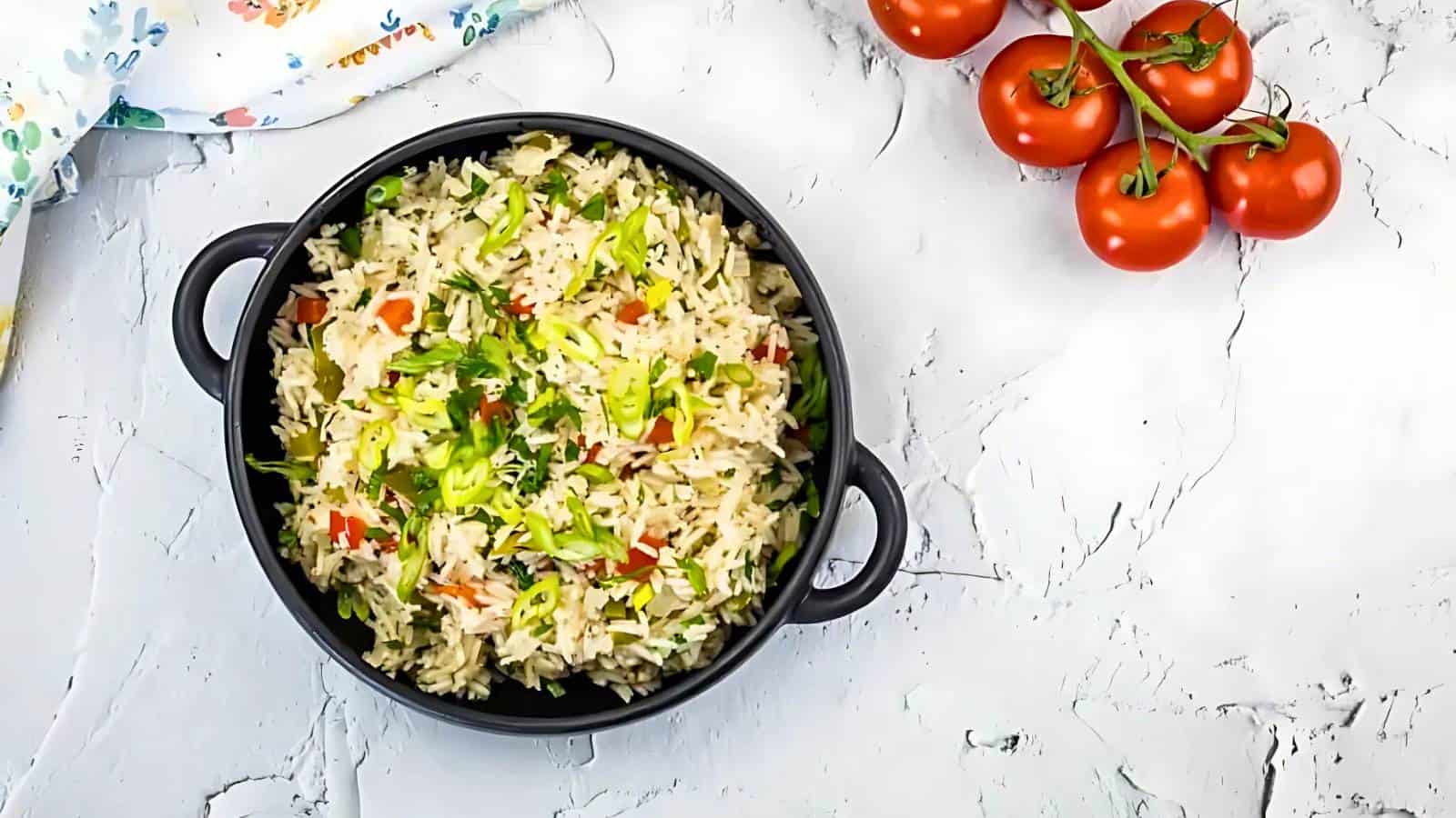 A black pot filled with cooked rice mixed with chopped vegetables sits on a white surface next to a cluster of vine tomatoes.