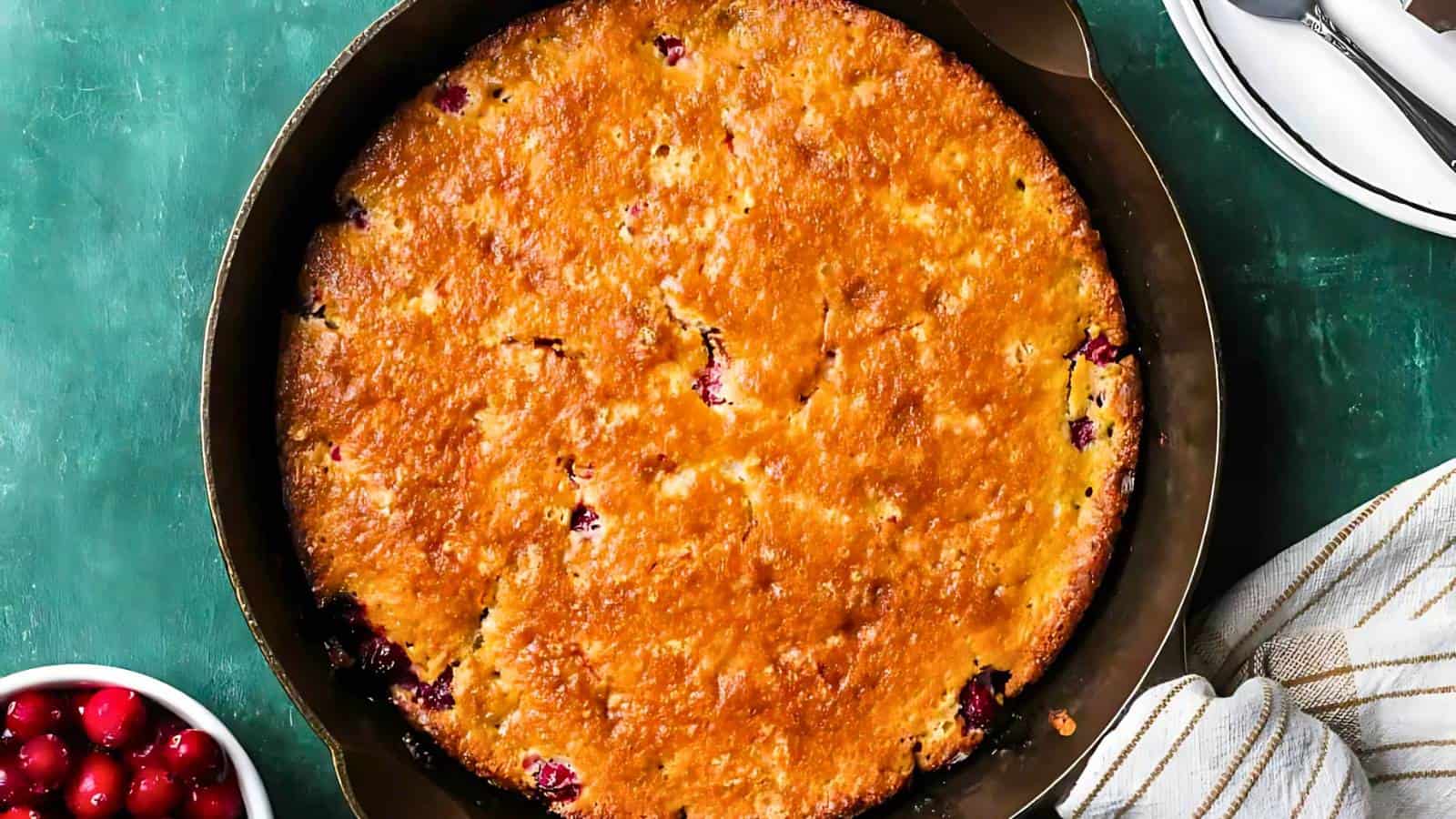 A golden-brown baked cranberry skillet cake sits in a round pan on a green surface, with a bowl of cranberries and a striped towel nearby.