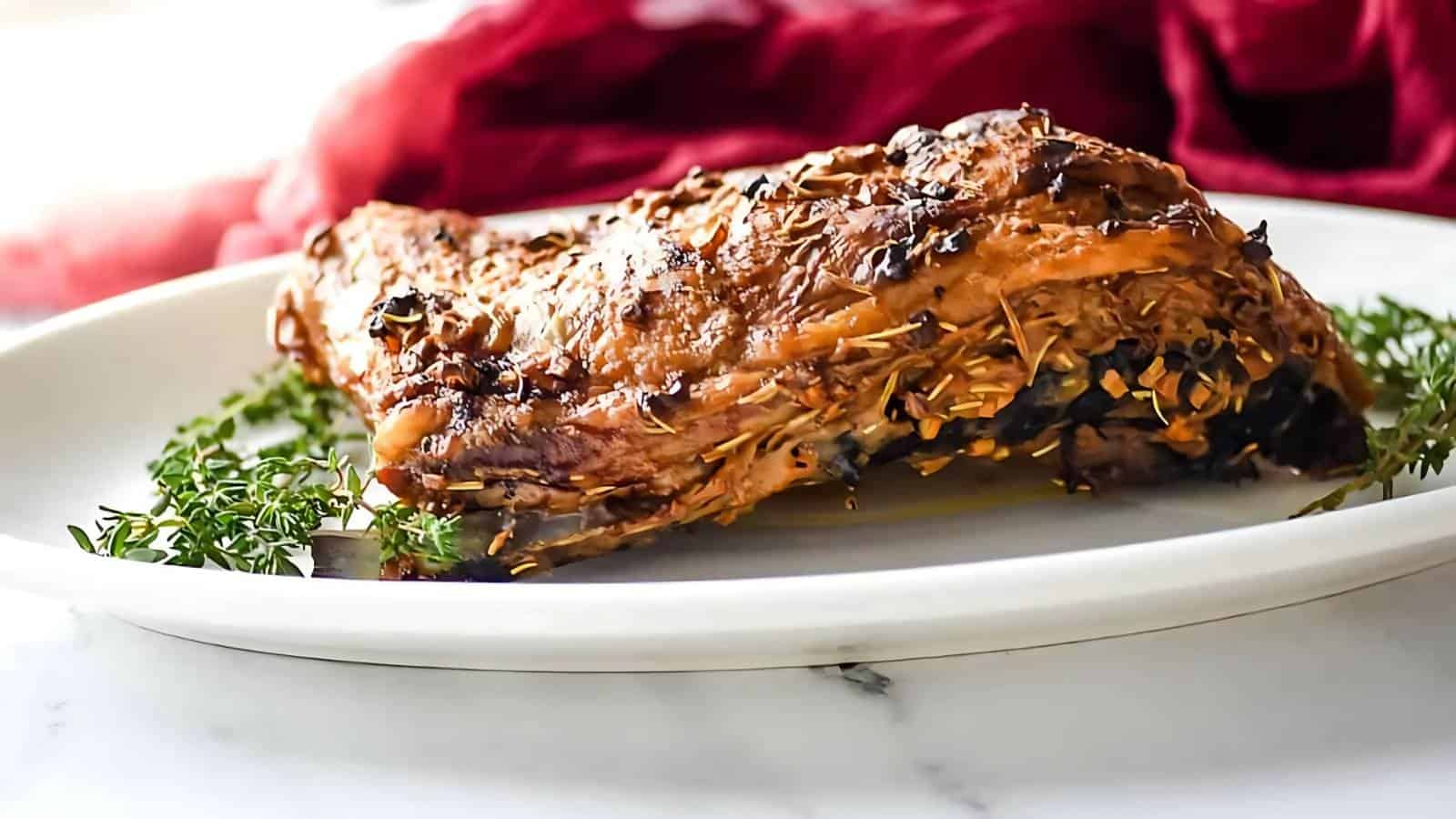 A cooked, herb-crusted rack of lamb sits on a white plate garnished with fresh sprigs of thyme, with a red cloth in the background.