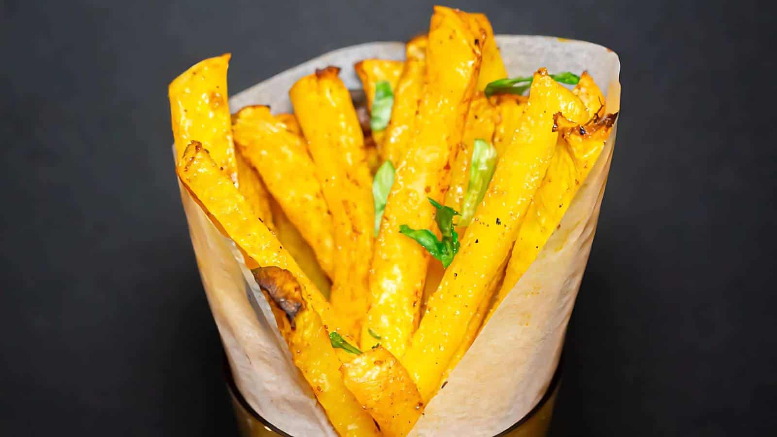A paper cone filled with seasoned, baked yellow fries garnished with chopped herbs, set against a plain dark background.