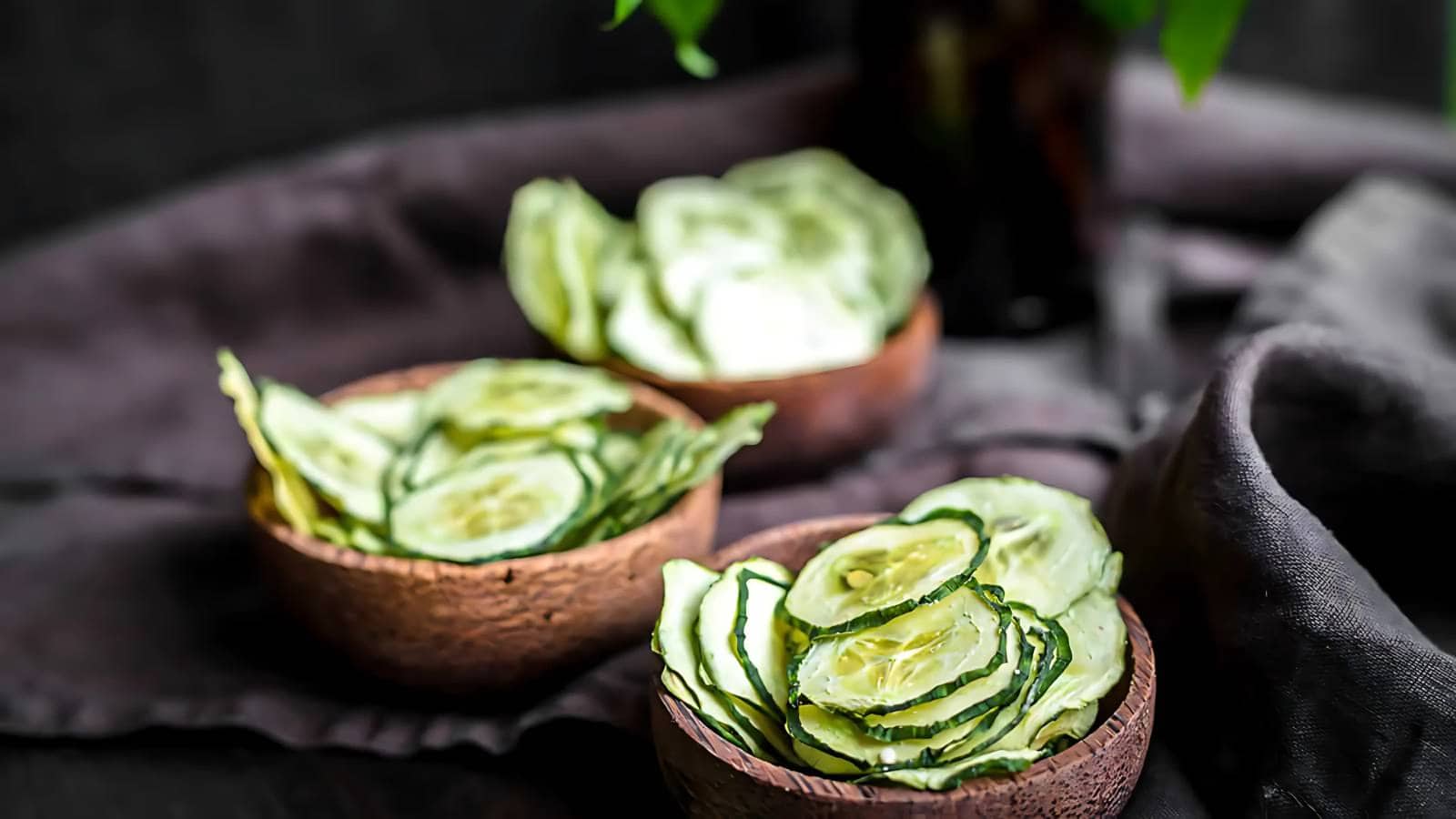 Three wooden bowls filled with thinly sliced cucumber chips are arranged on a dark cloth surface, with a blurred background.