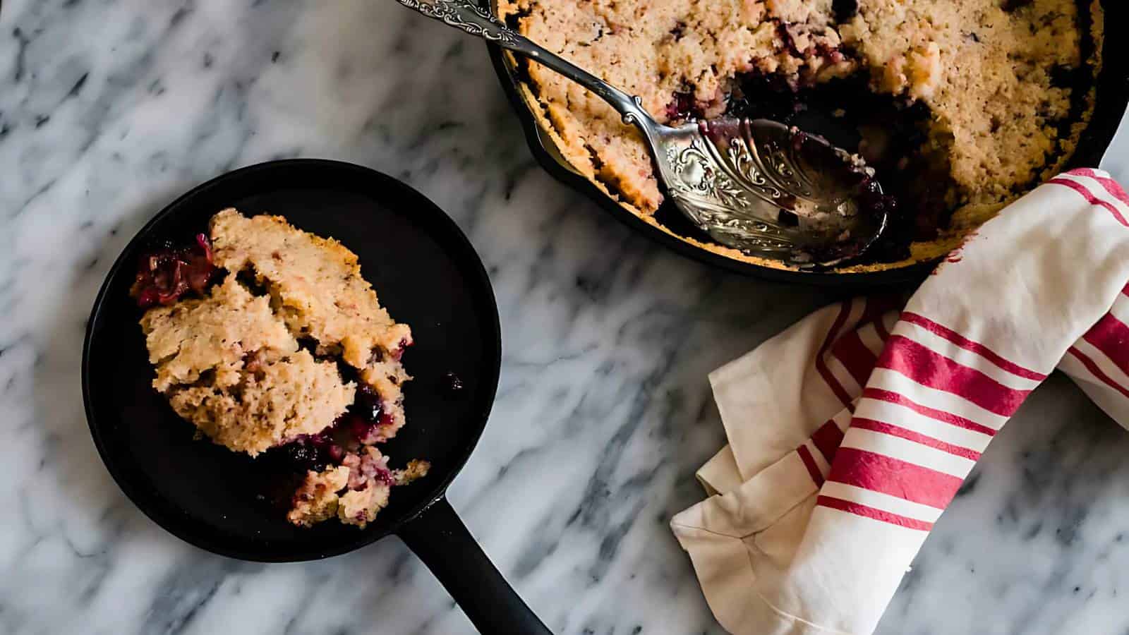A serving of berry cobbler on a black plate sits next to a cast iron skillet containing the remainder of the cobbler, with a metal serving spoon and a striped cloth on a marble surface.