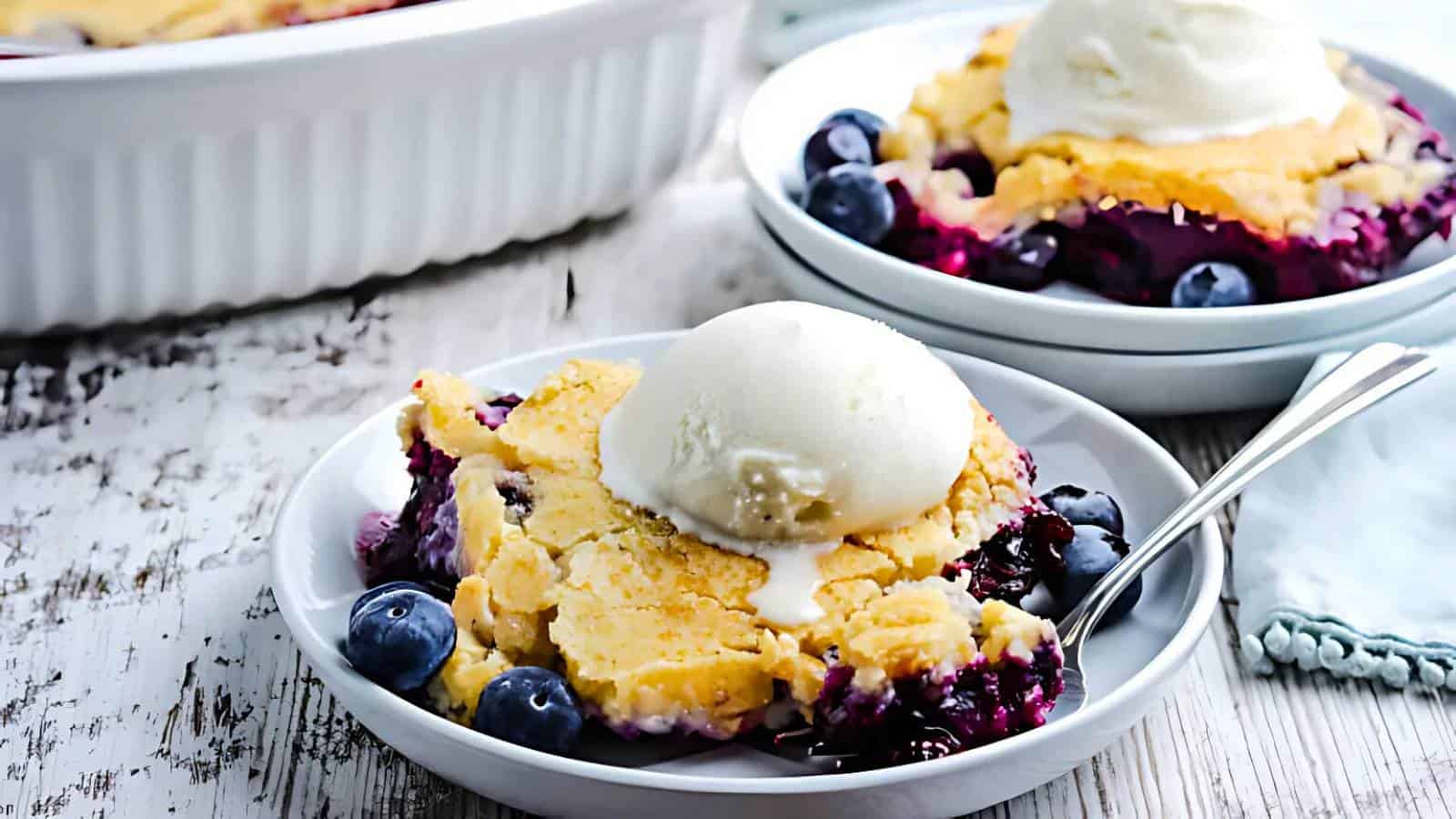 A serving of blueberry cobbler topped with a scoop of vanilla ice cream on a white plate, garnished with fresh blueberries.