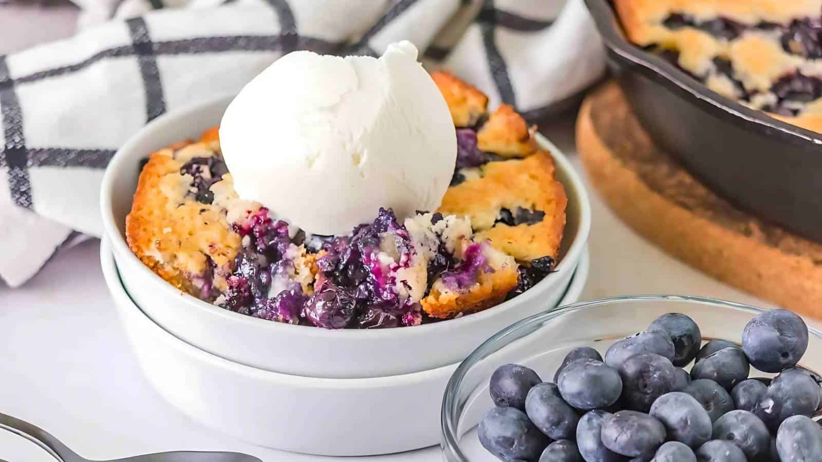 A bowl of blueberry cobbler topped with a scoop of vanilla ice cream, next to a bowl of fresh blueberries and a cast iron skillet with more cobbler.