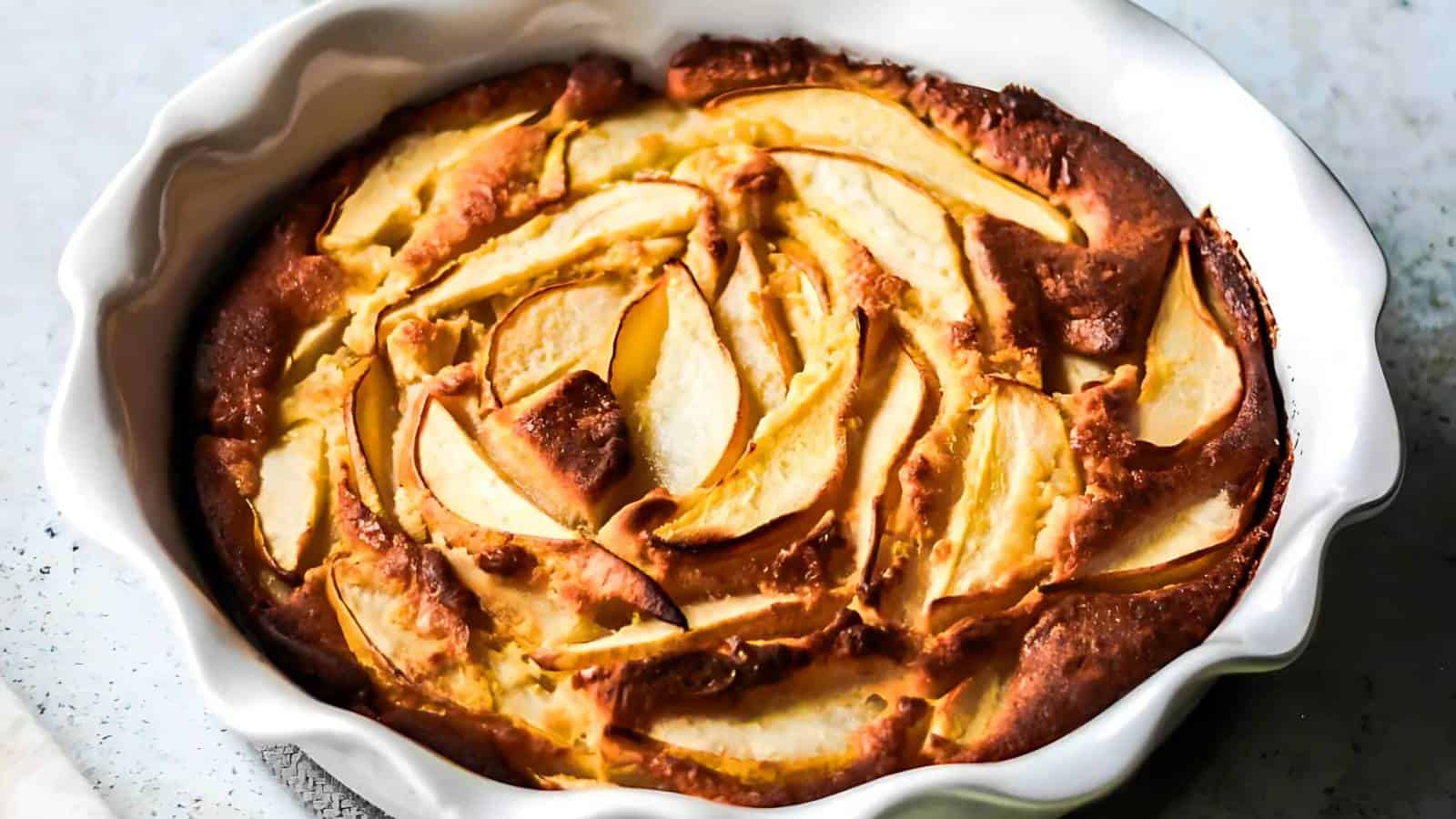 A baked apple clafoutis with golden brown edges and arranged apple slices in a white scalloped ceramic dish.