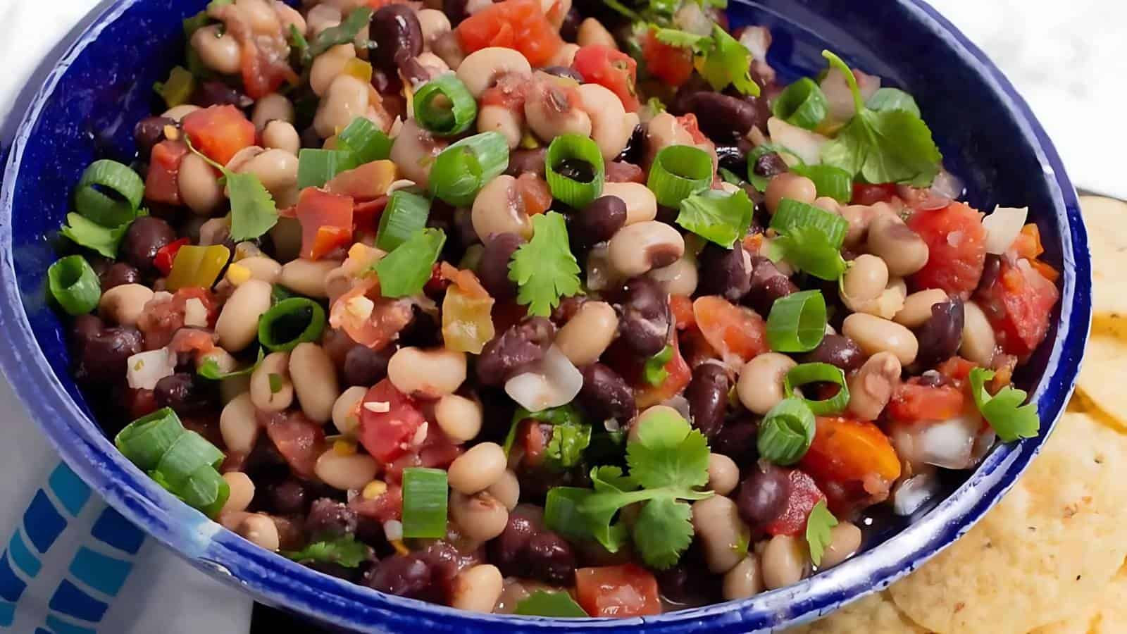 A blue bowl filled with a bean salad made of black beans, black-eyed peas, chopped tomatoes, onions, cilantro, and green onions, with tortilla chips beside it.