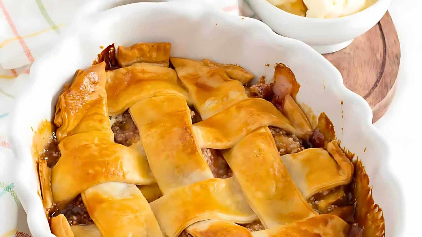 A baked pie with a golden-brown lattice crust is in a white dish, with a bowl of vanilla ice cream on a wooden coaster in the background.