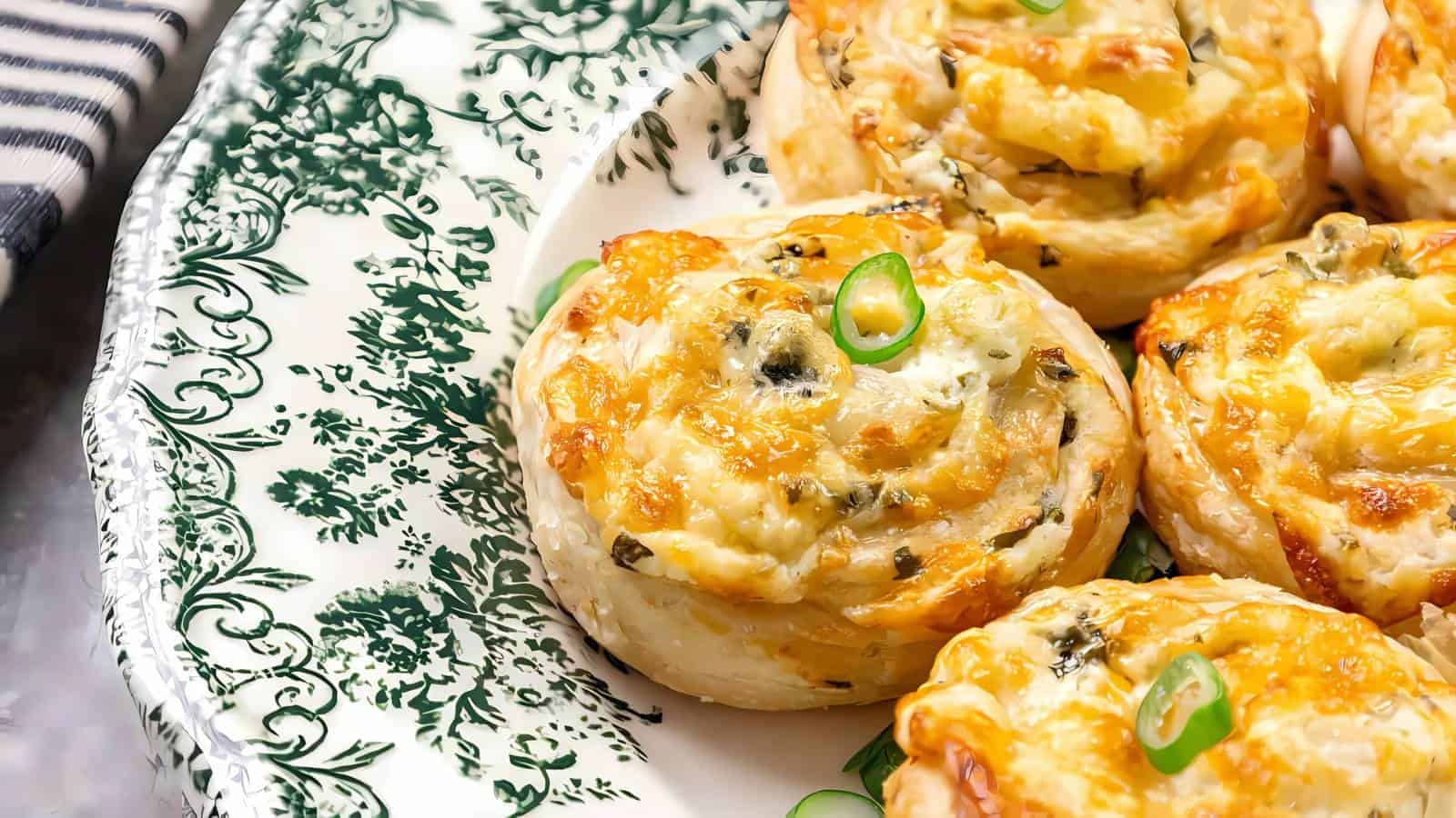 A decorative plate holds several golden-brown, cheese and herb pastry pinwheels, garnished with sliced green onions.