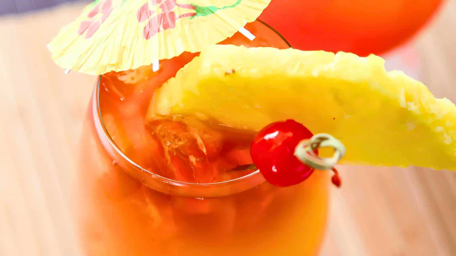 A close-up of a tropical cocktail with ice, garnished with a pineapple slice, a cherry, and a paper umbrella.