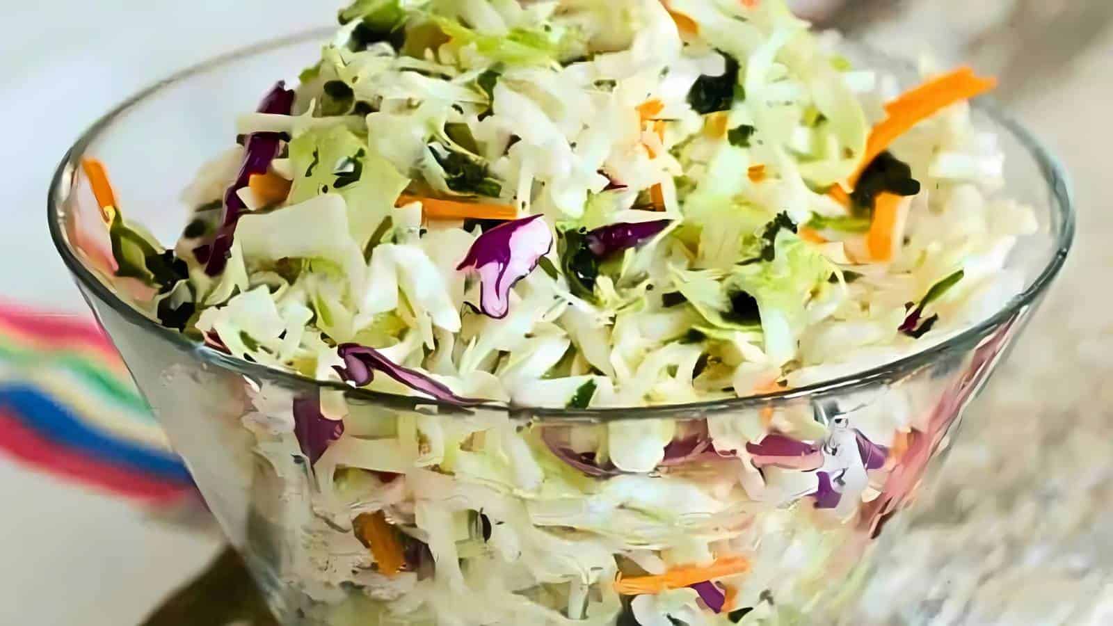 A glass bowl filled with coleslaw made of shredded cabbage, carrots, and purple cabbage on a light surface.