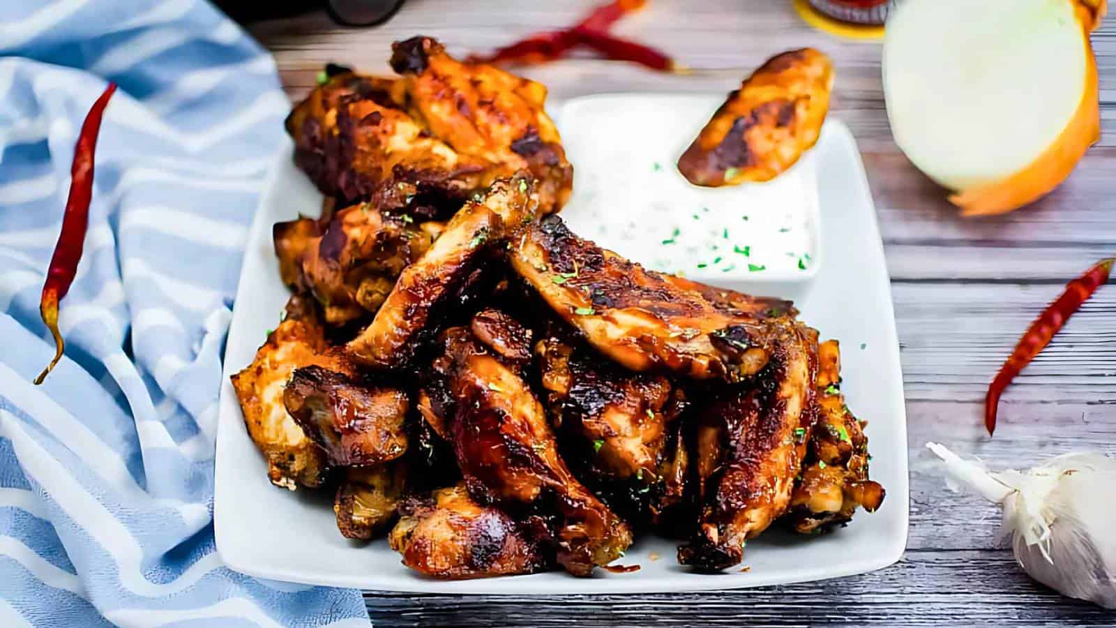 A plate of grilled chicken wings with a side of dipping sauce, garnished with herbs, on a wooden table with chili peppers, garlic, and an onion nearby.