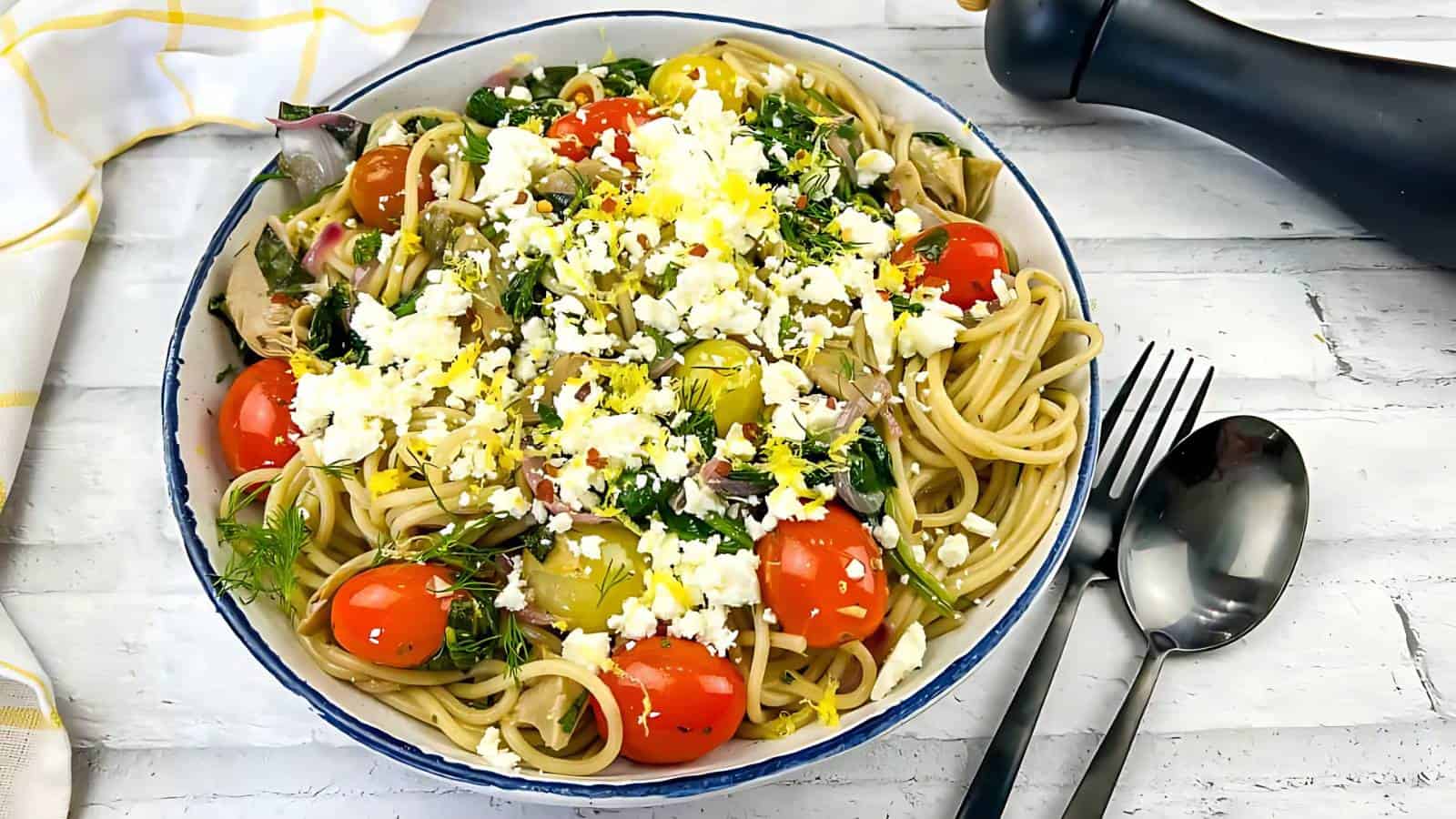 A bowl of spaghetti topped with cherry tomatoes, greens, crumbled cheese, and herbs, with a fork and spoon on the side and a pepper grinder in the background.