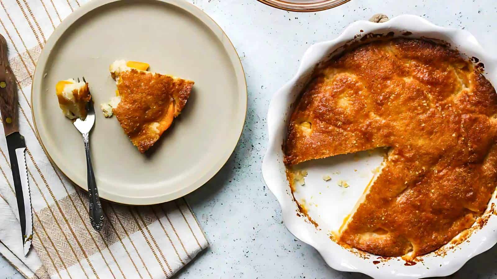 A round baking dish with a golden-brown cobbler, one slice removed and placed on a plate beside it; a fork rests on the plate near the slice.