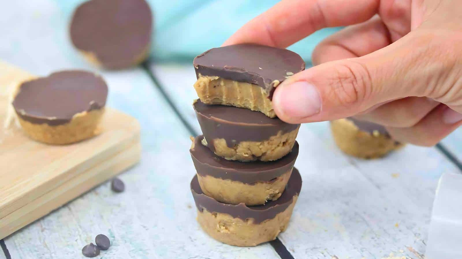 A hand holds a stack of three chocolate and peanut butter cups, with a bite taken from the top one; more cups and chocolate chips are in the background.