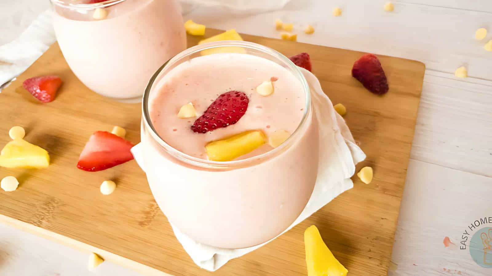 A glass of pink smoothie topped with a strawberry slice, pineapple chunks, and white chocolate chips on a wooden board with scattered fruit pieces.